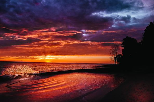 A wave crashes as a dramatic sunset covers the sky.